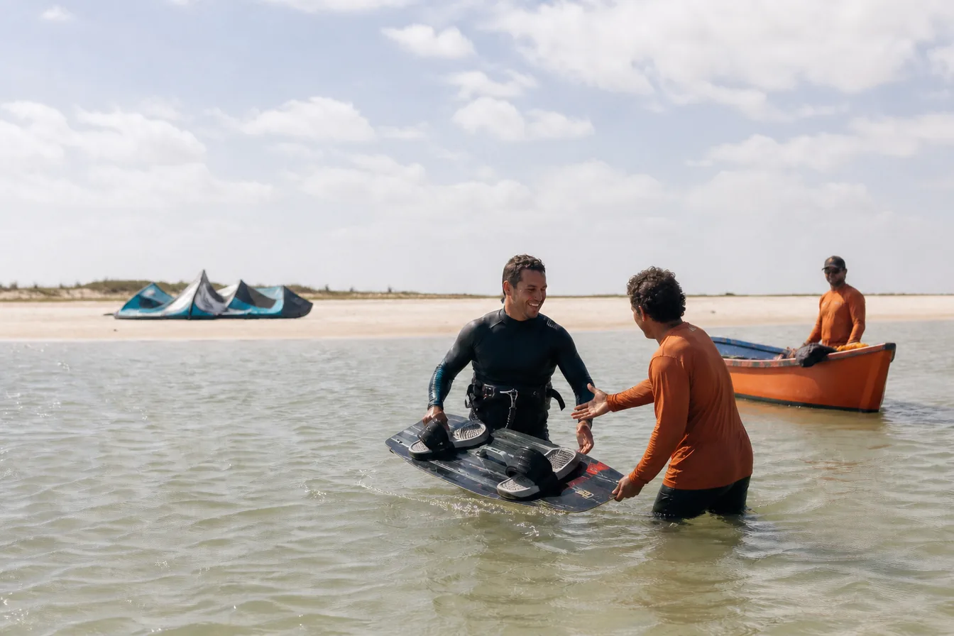 Velejador voltando da água com a prancha enquanto um integrante da equipe o ajuda, com barco de apoio ao fundo