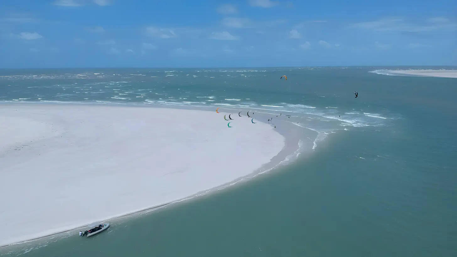 Vista aérea de um banco de areia branca contornado por água verde-turquesa, com vários kitesurfistas velejando e um barco de apoio na orla